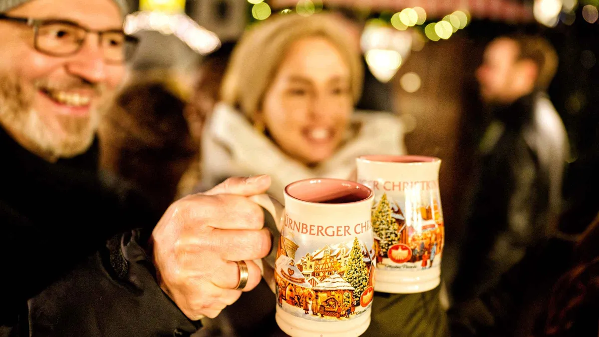 Two travelers raising Christkindlesmarkt mugs of mulled wine at an evening market stall